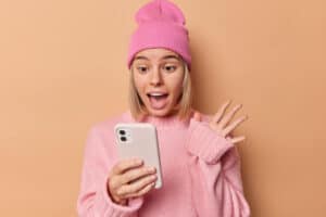 une adolescente portant un pull et un bonnet rose clair semble surprise en regardant son smartphone