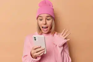 une adolescente portant un pull et un bonnet rose clair semble surprise en regardant son smartphone