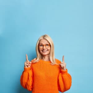 une femme, portant un pull orange,  lèves les deux index et semble heureuse 
